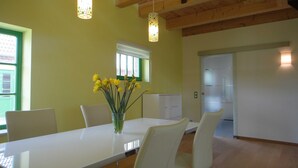 A dining area with a white table, chairs, and a vase of yellow flowers. There is a window with green trim and a door with frosted glass panels.