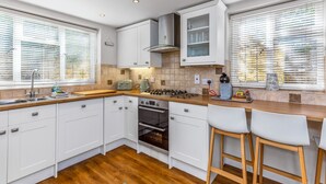 Cocina moderna con armarios blancos, encimera de madera, estufa a gas y horno empotrado. Hay dos taburetes de bar y una ventana con persianas.