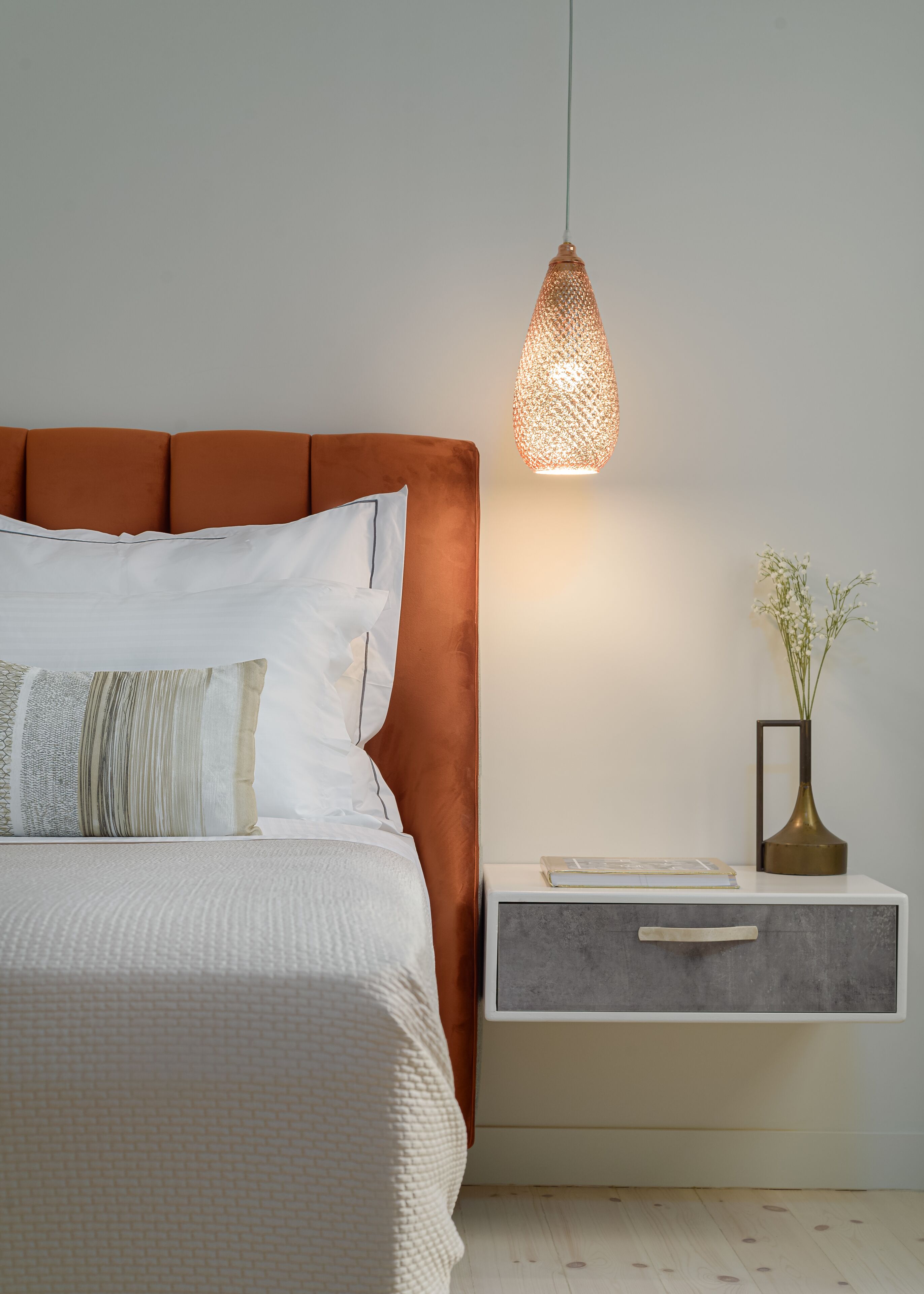 A neatly made bed with a brown headboard, a bedside table with a vase of flowers, and a hanging light fixture.