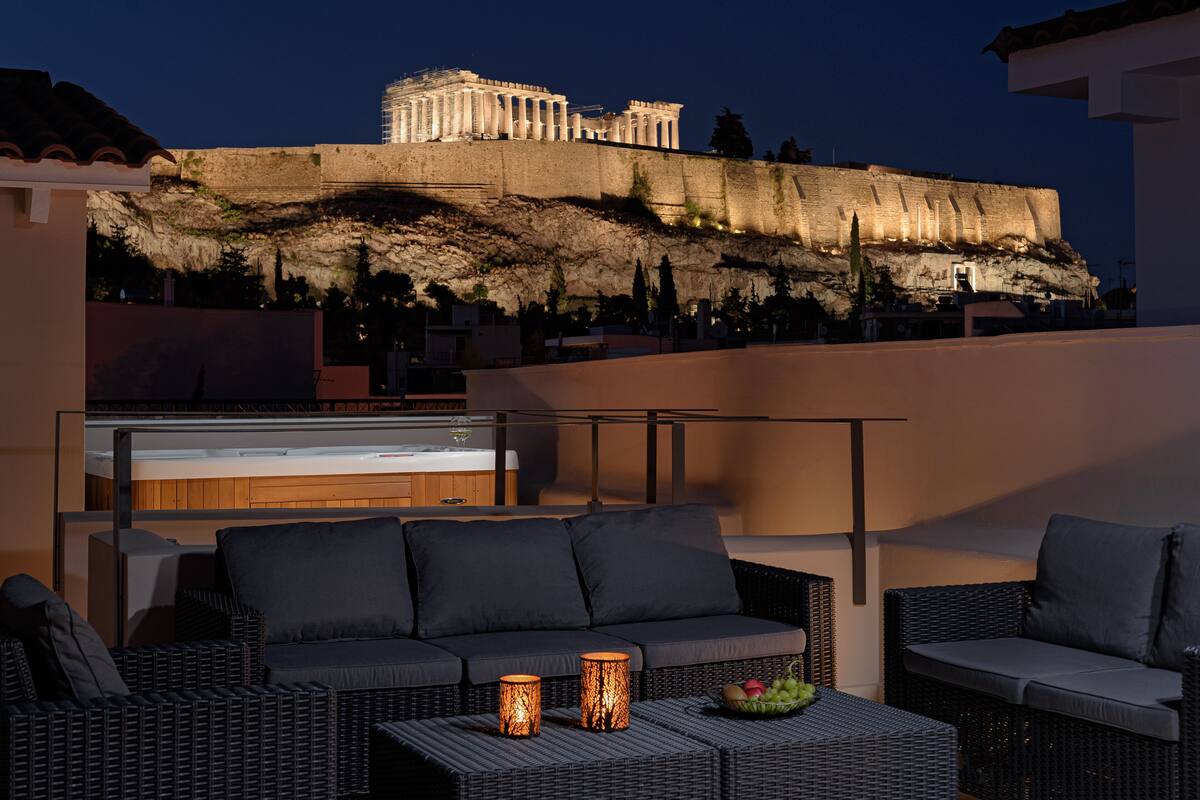 A rooftop terrace with wicker furniture, a bar, and a view of the Acropolis at night.