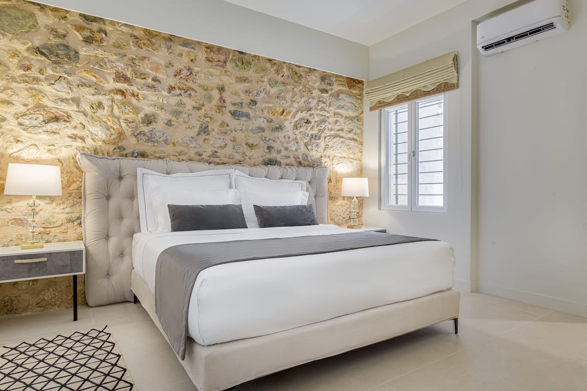 A bedroom with a stone wall, a large bed with white bedding, two bedside tables with lamps, and a window with blinds.