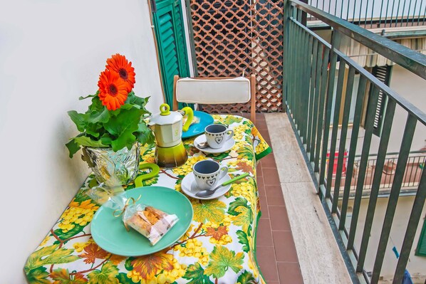 A balcony with a table set for two, featuring a floral tablecloth, a green chair, a vase with a single flower, and a green plate with food.