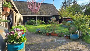 Un giardino con diverse piante in vaso e fiori, una recinzione in legno e un edificio con tetto di paglia sullo sfondo.