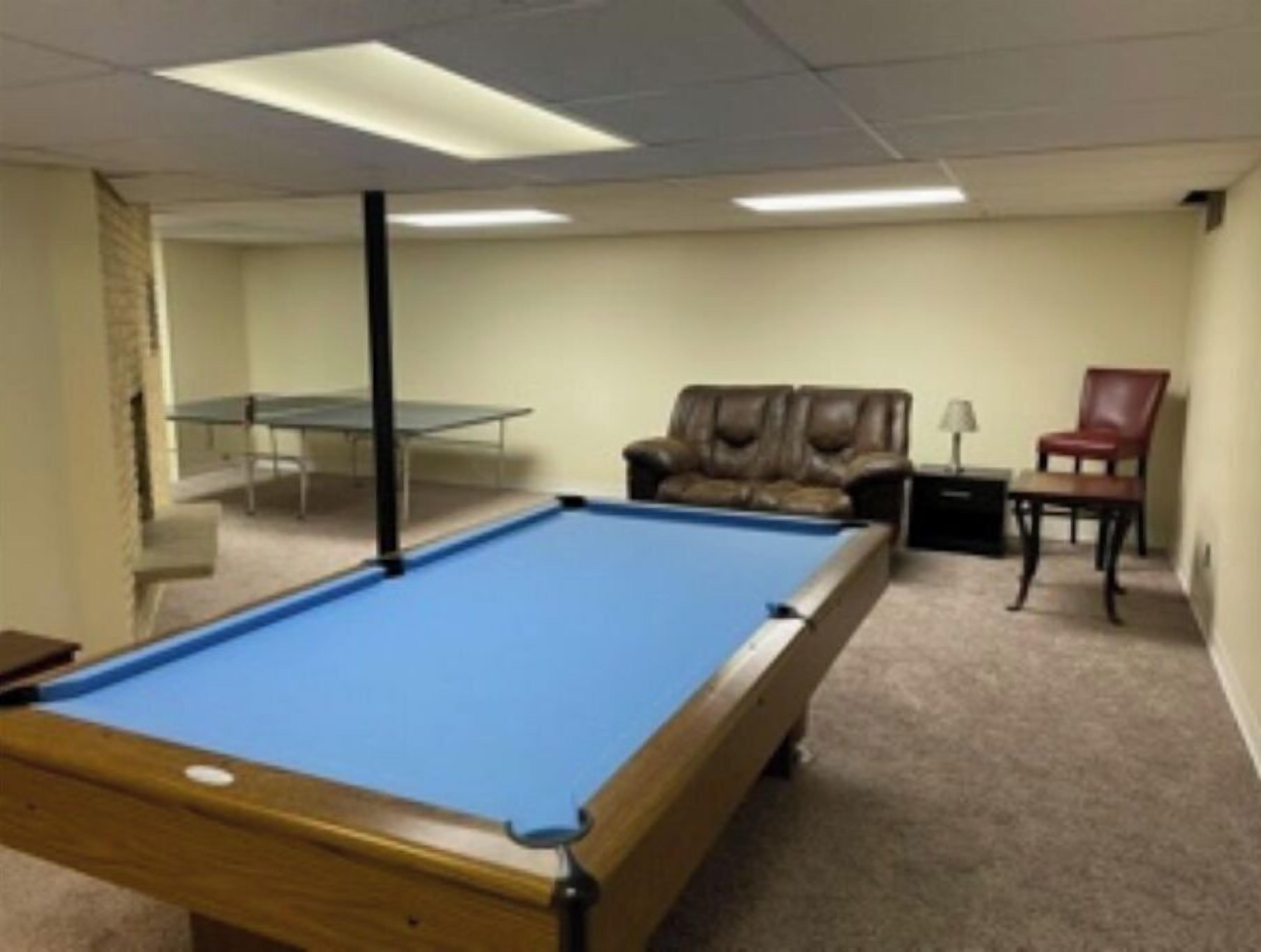 Une table de billard dans une pièce dont le sol est recouvert d’une moquette, avec un canapé en cuir, un fauteuil rouge et une lampe de table.