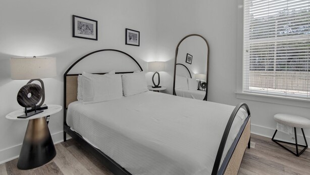 A modern bedroom with a large bed, two bedside tables with lamps, a round side table, and a window with blinds. - Sea It All - 2303626 (Santa Rosa Beach)