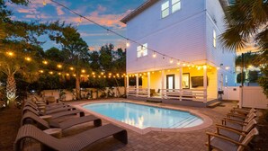 A pool area with lounge chairs, a white building with a porch, and string lights. - Sea It All - 2303626 (Santa Rosa Beach)
