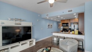 A modern living room with a flat-screen TV, a ceiling fan, and a kitchen area with wooden cabinets and a bar stool. - Sterling Breeze 403 - 2180902 (Panama City Beach)