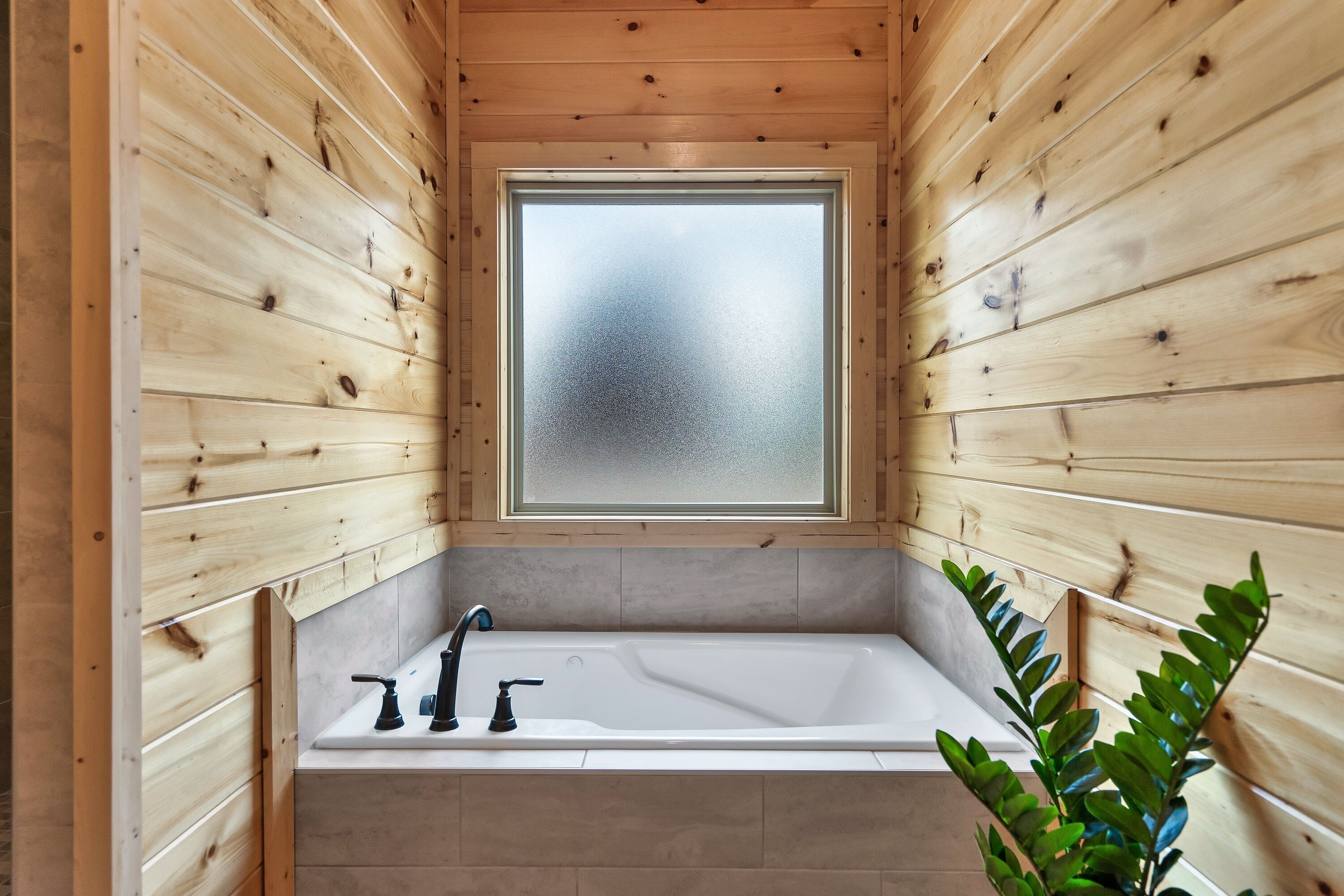A wooden cabin bathroom with a large window, a bathtub, and a plant.
