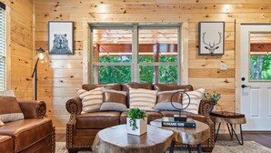 Een gezellig chaletinterieur met een bruine leren bank, twee ronde houten tafels en een groot raam met uitzicht op groen.