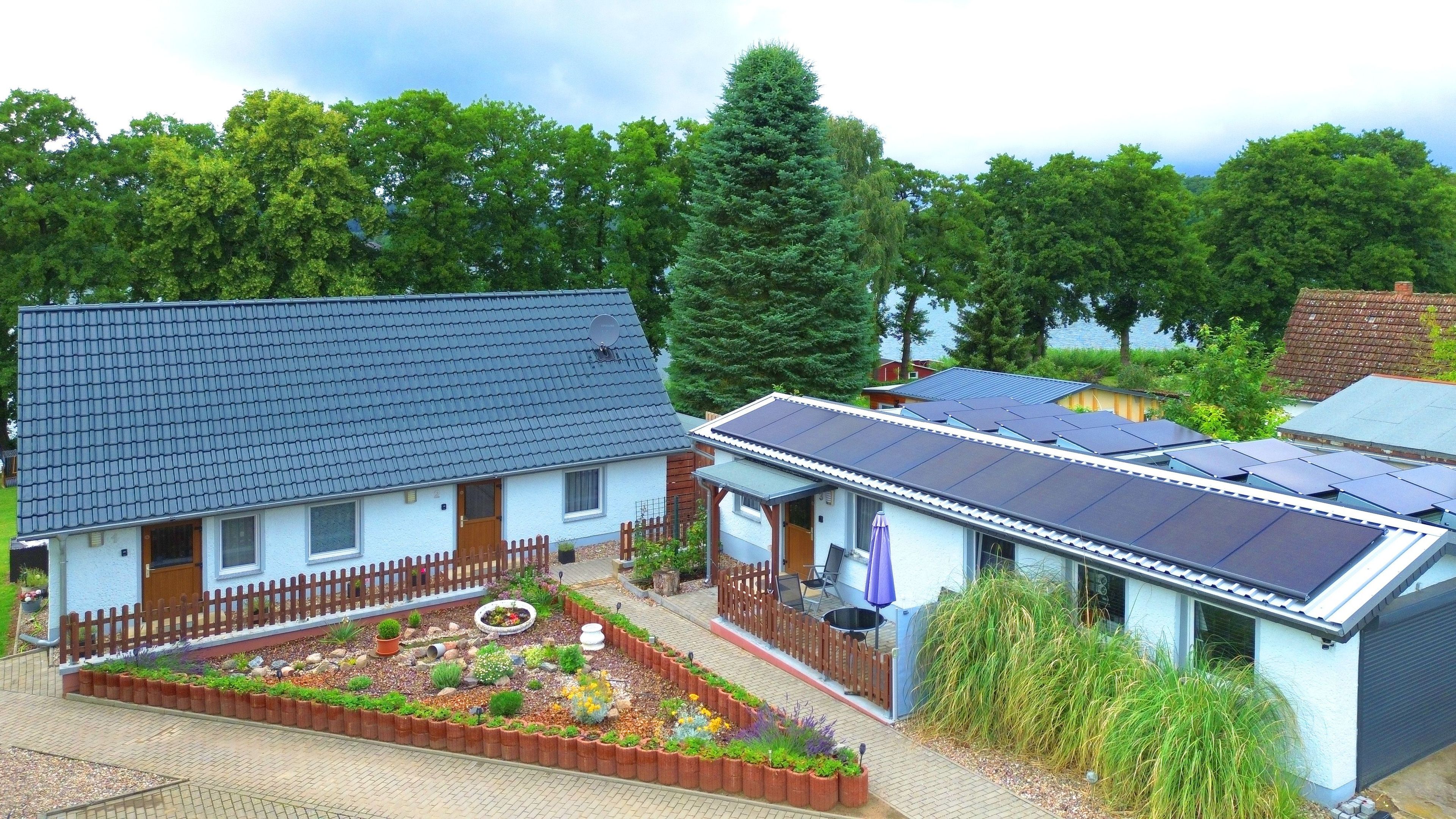 Una casa con techo de paneles solares, jardín cercado y un árbol grande al fondo.