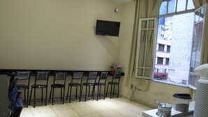 Una habitación con un televisor montado en la pared, una ventana con cortinas y una fila de sillas contra la pared.