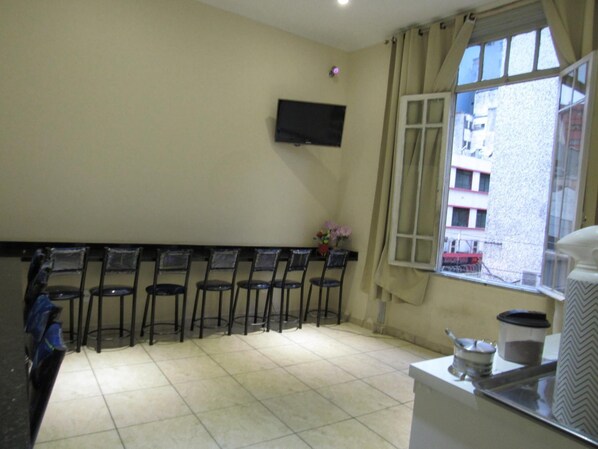 A room with a TV mounted on the wall, a window with curtains, and a row of chairs against the wall.