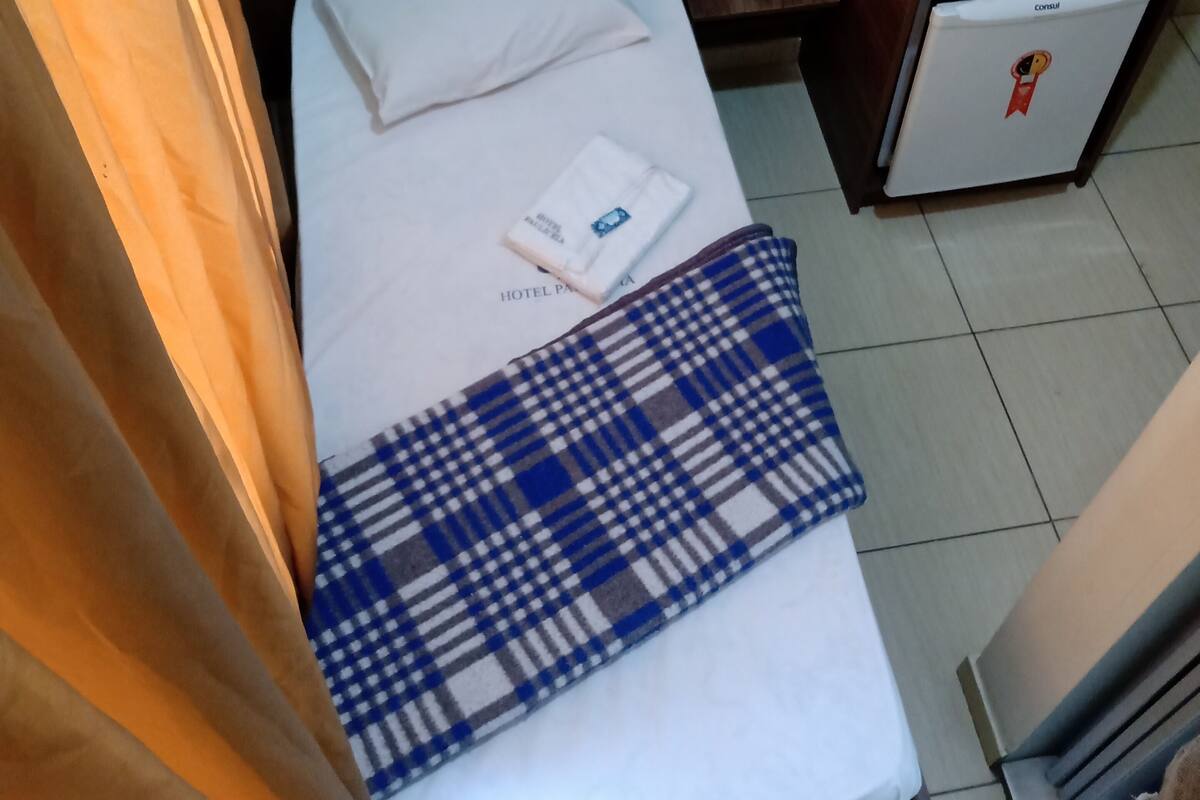 Pieni hotellihuone, jossa on yksi sänky, puinen vaatekaappi, pieni pöytä, jossa on kaukosäädin ja rulla WC-paperia, sekä minijääkaappi.