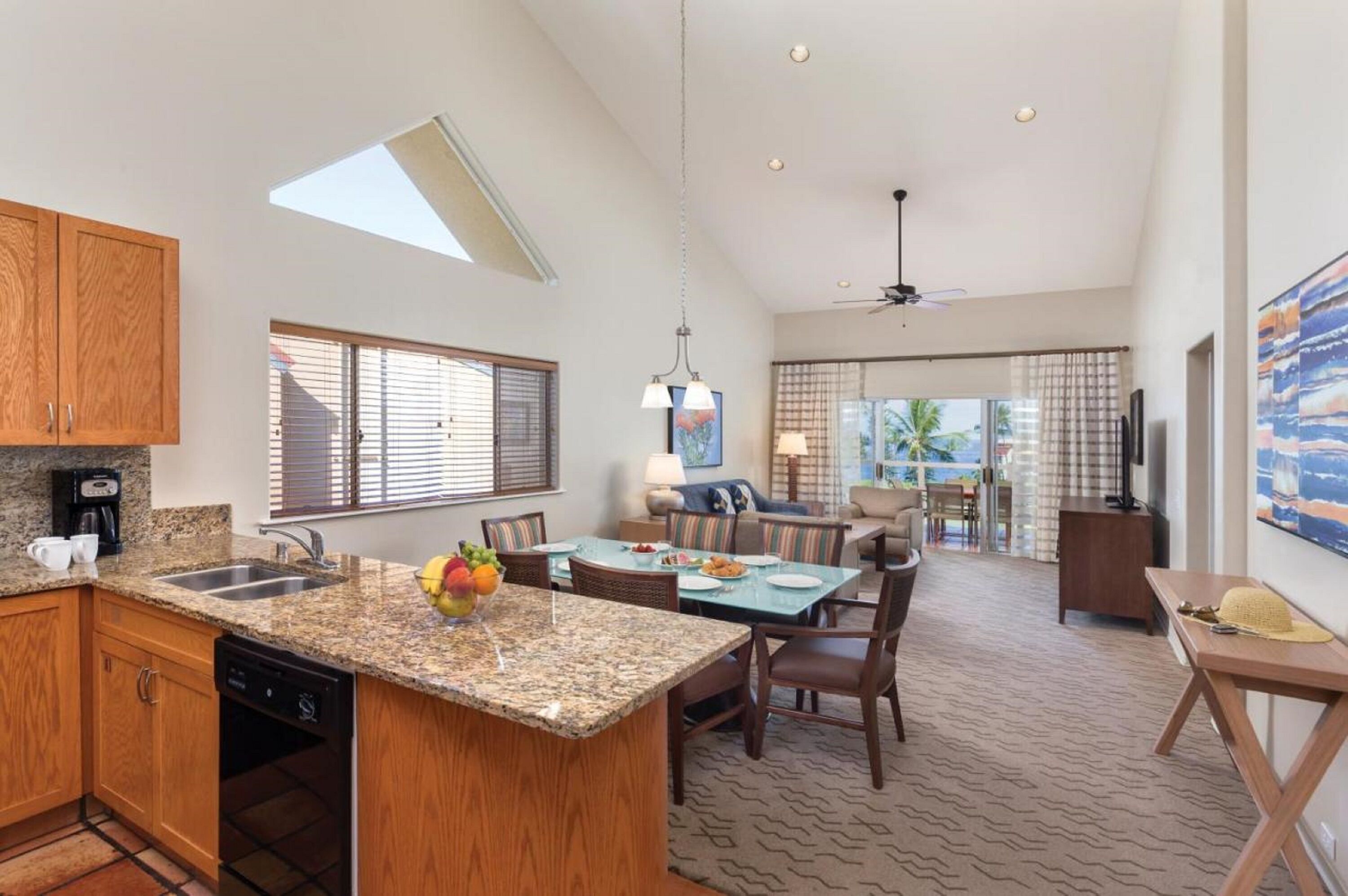 A spacious kitchen with granite countertops, wooden cabinets, and a built-in dishwasher. There is a dining area with a table and chairs, and a living room with a sofa and a TV.