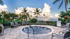 A circular hot tub surrounded by lounge chairs, with a stone patio and palm trees in the background.