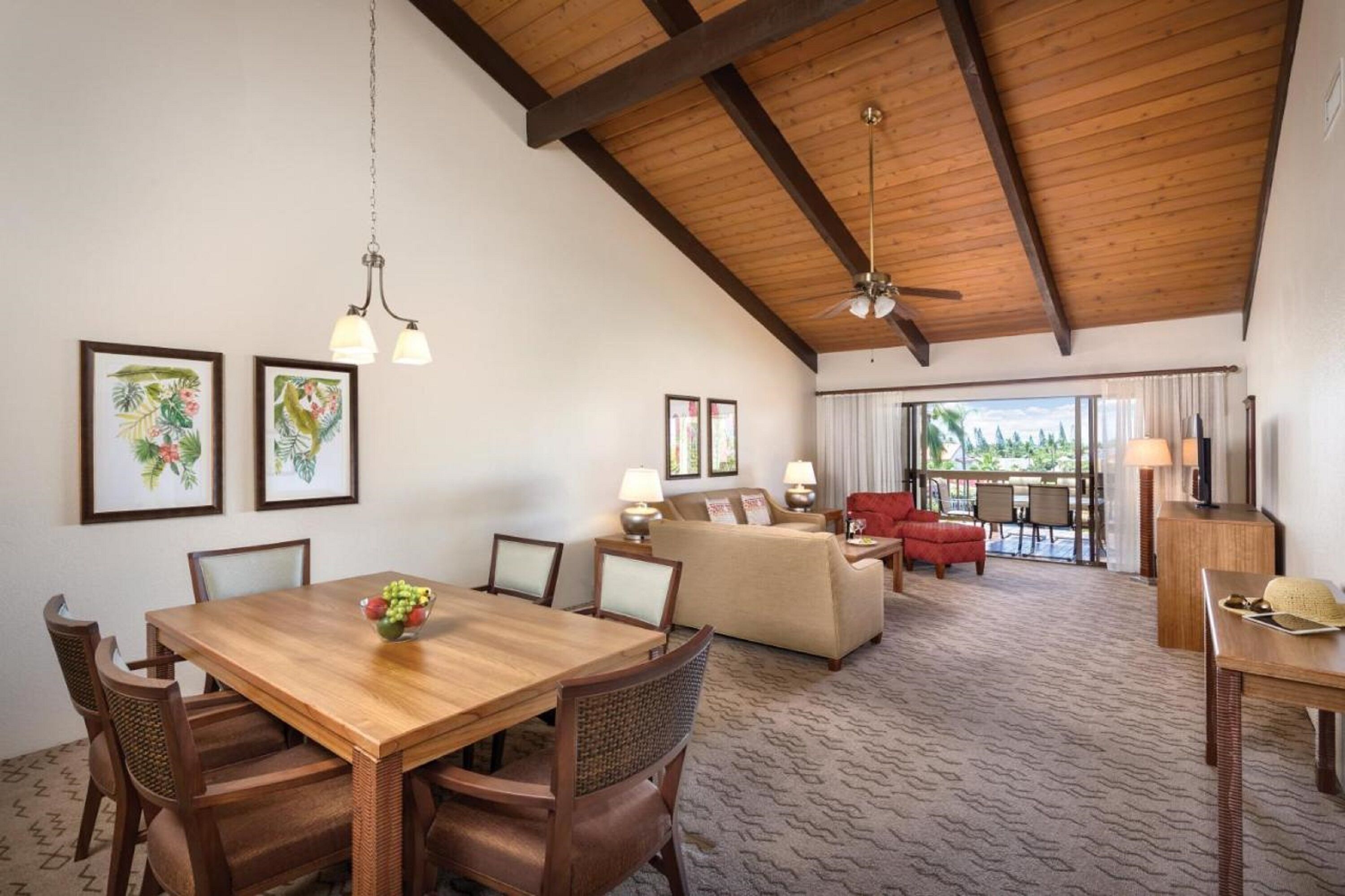 A spacious room with a wooden dining table, chairs, a sofa, and a red armchair. There are framed pictures on the wall, a ceiling fan, and a view of the outdoors through large windows.