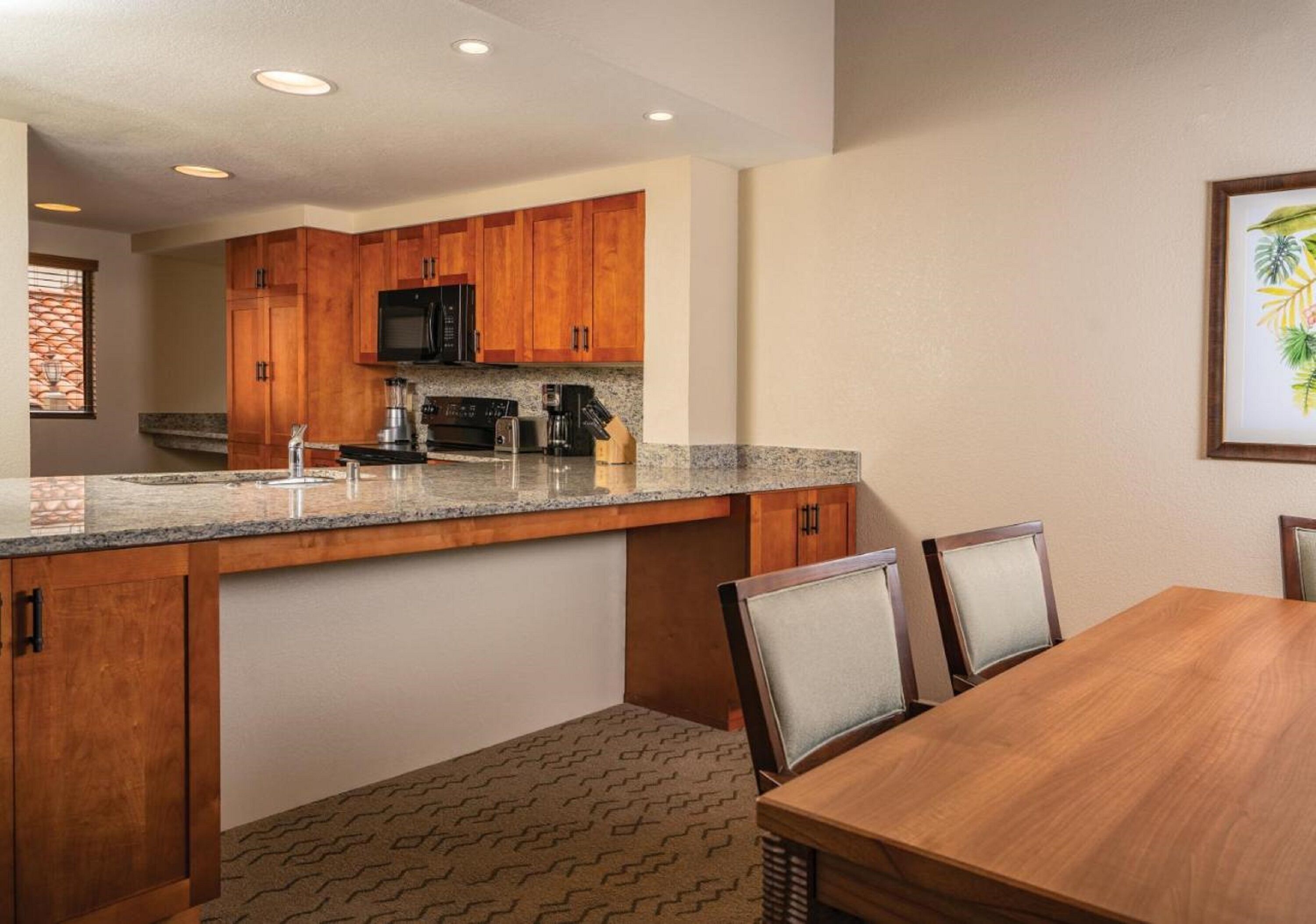 A kitchen with wooden cabinets, a granite countertop, a microwave, and a coffee maker. There is a dining table with chairs.