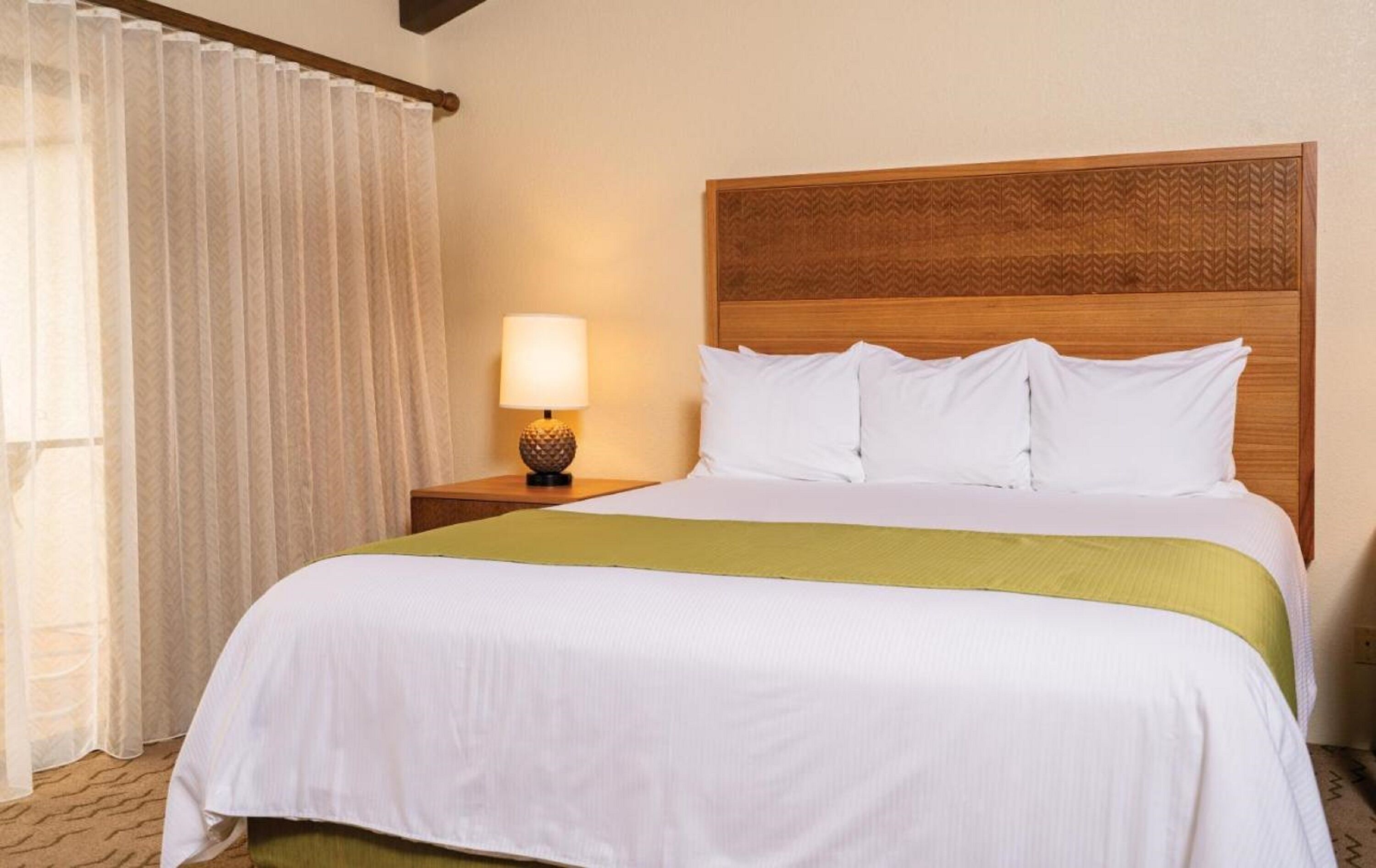 Une chambre d’hôtel avec un grand lit, une tête de lit en bois, une table de chevet avec une lampe et des rideaux légers.