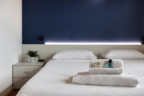 A neatly made bed with a folded towel and toiletries, a bedside plant, and a simple bedside table.