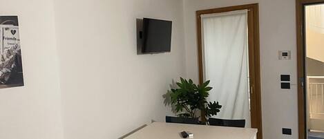 A dining area with a white table and chairs, a potted plant, a TV on the wall, and a door with a curtain.