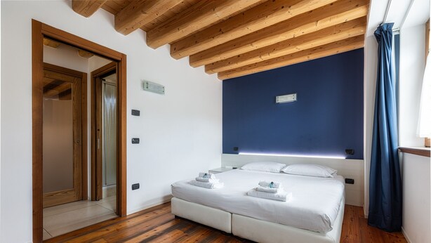 A bedroom with a wooden ceiling, a bed with white linens, a blue accent wall, and a wooden floor.