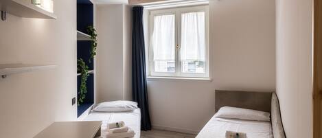 A small, well-lit room with two beds, a desk, and a window with curtains.