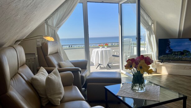 A modern living room with a leather sofa, a glass coffee table with a vase of flowers, a flat-screen TV, and a balcony with a view of the sea. - N36 (BC.a5) - Stranddomicil Steiner (Büsum)