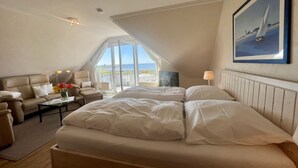 A bedroom with a large bed, a sofa, a chair, a table with a vase of flowers, and a large window offering a view of the sea. - N36 (BC.a5) - Stranddomicil Steiner (Büsum)