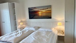 Een slaapkamer met een bed, twee bedlampen, een schilderij van een zonsondergang en een witte kledingkast.