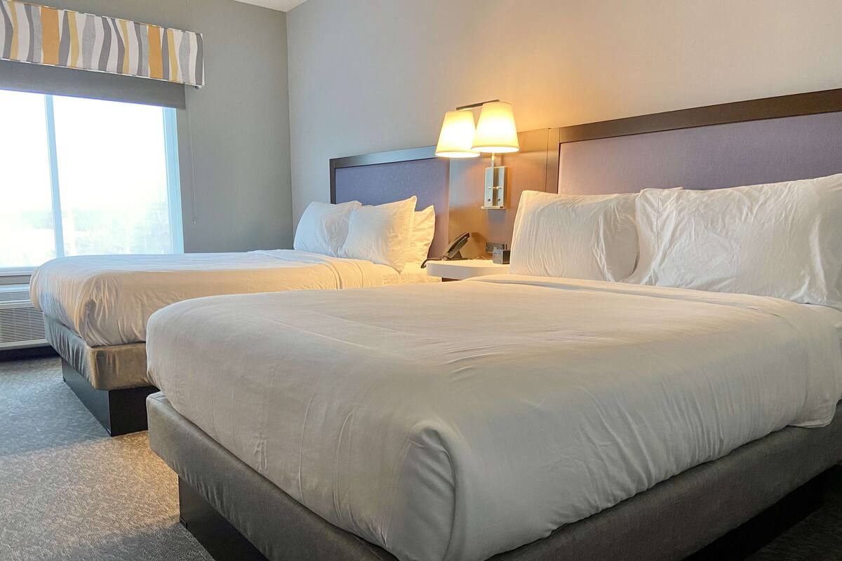 Hotellihuone, jossa on kaksi sänkyä, yöpöytä lampun kanssa ja ikkuna, jossa on sälekaihtimet.