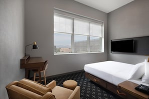 Habitación de hotel con cama, escritorio, silla, sofá, televisor y ventana con vista a la ciudad.