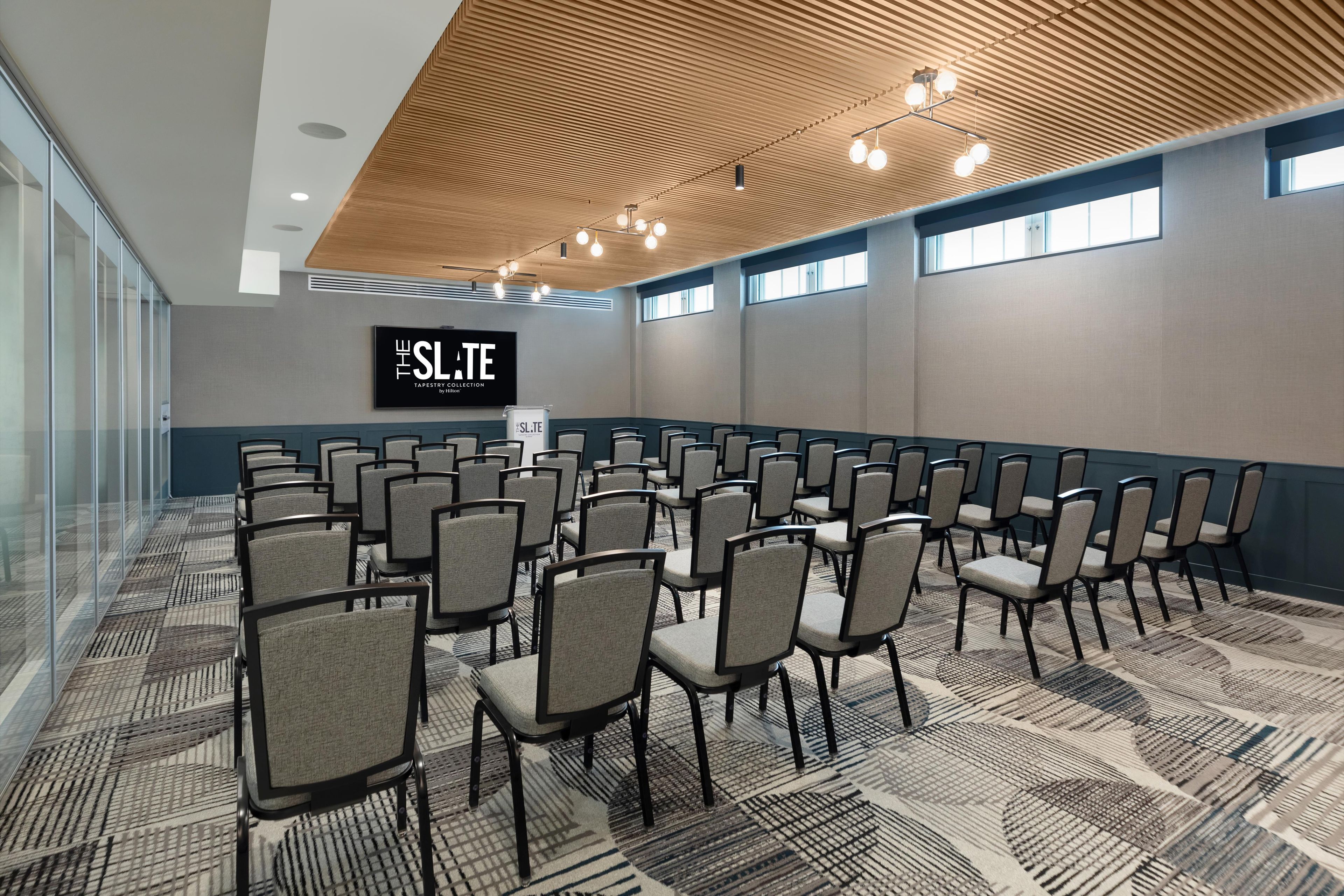 A conference room with rows of chairs, a wooden ceiling, and a large screen displaying 'THE SLATE TAPESTRY COLLECTION by Hilton'.