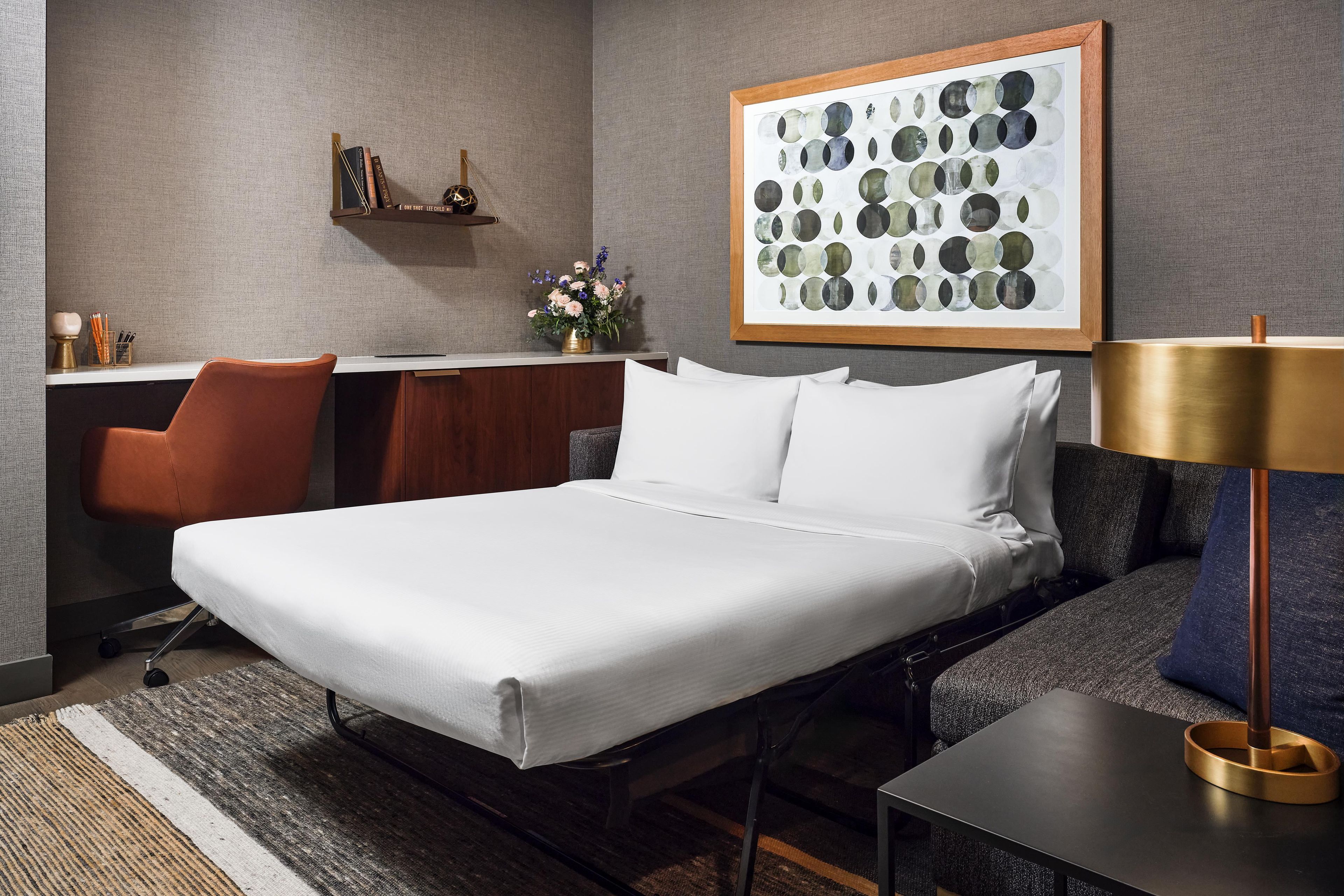 Una habitación de hotel con cama, escritorio, sillón, lámpara y una obra de arte en la pared.