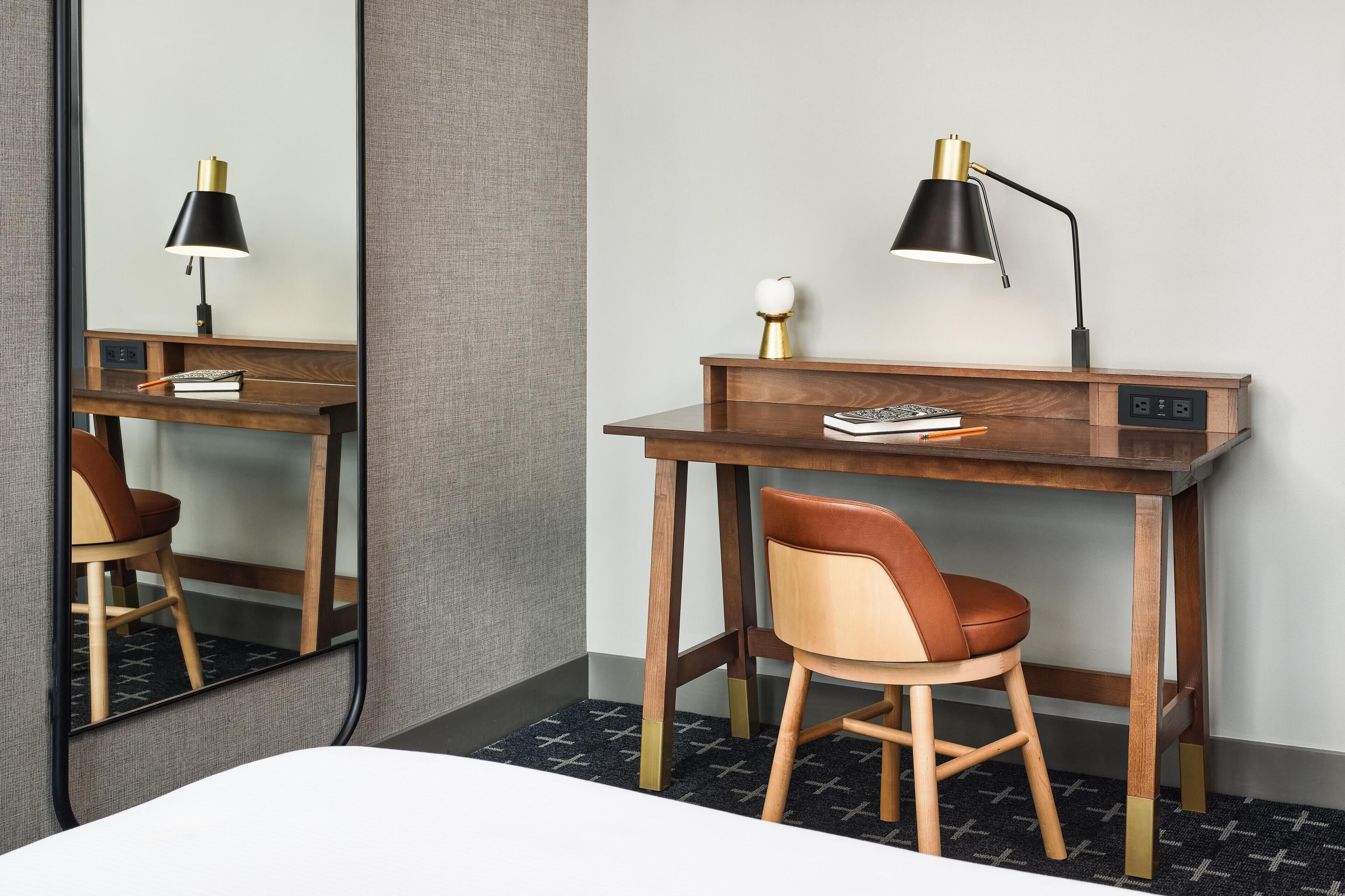 Une chambre d’hôtel avec un bureau en bois, une chaise, une lampe de bureau, un miroir et un petit objet décoratif posé sur le bureau.