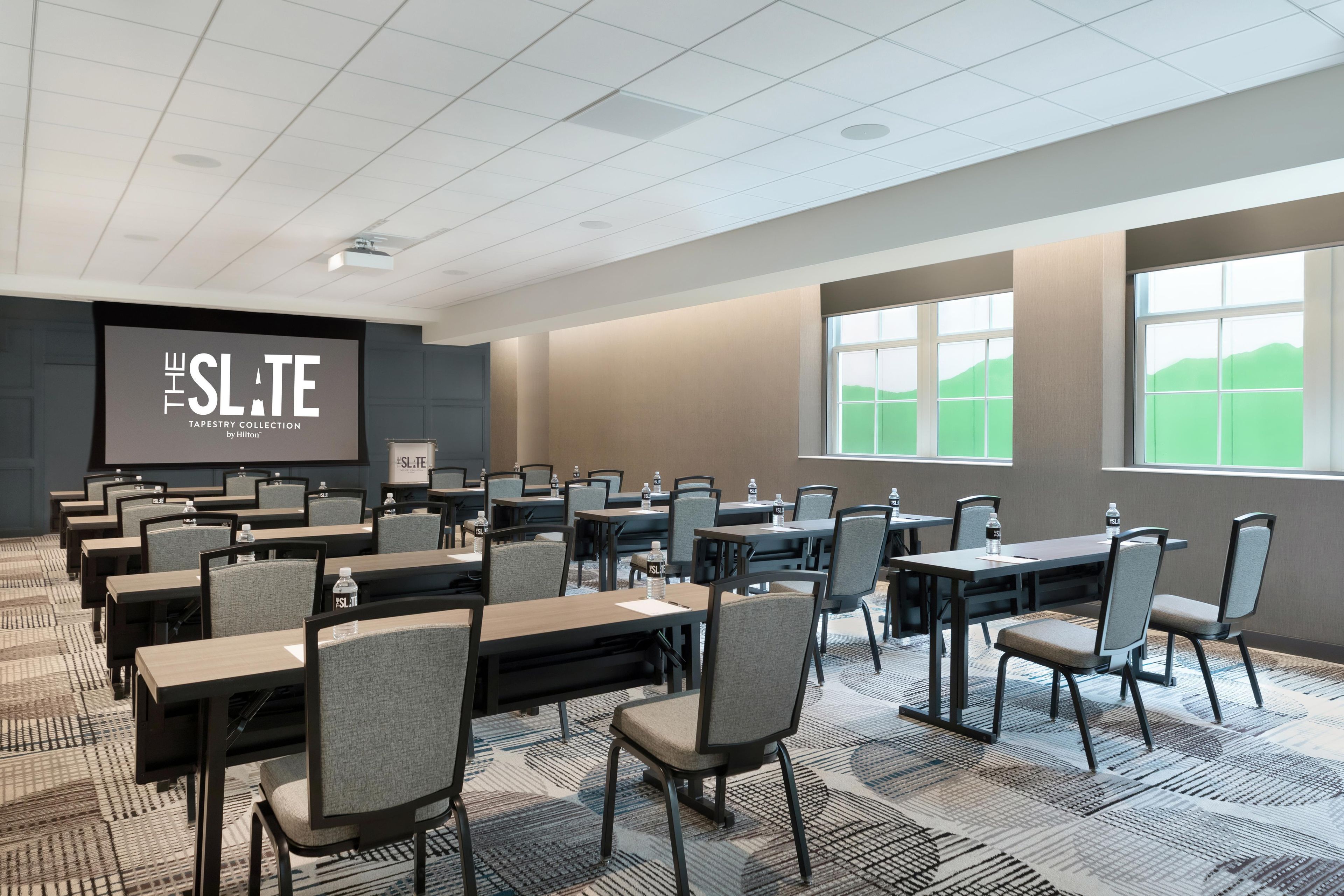 A conference room with multiple tables and chairs, a large screen displaying 'THE SLATE TAPESTRY COLLECTION by Hilton', and large windows with a view of green hills.