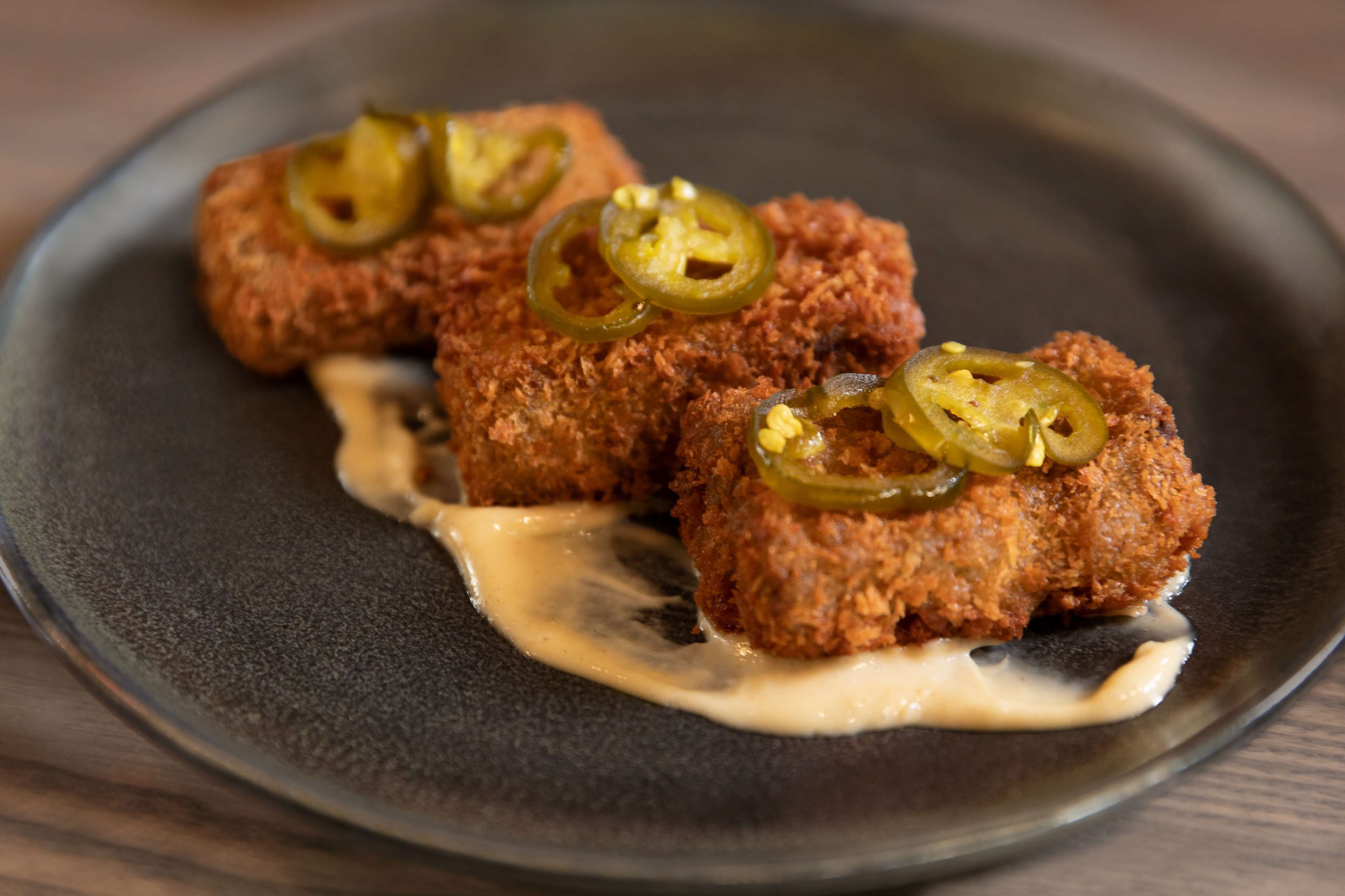 Trois morceaux de nourriture panés et frits, garnis de tranches de piment jalapeño, présentés sur une assiette sombre avec une sauce crémeuse.