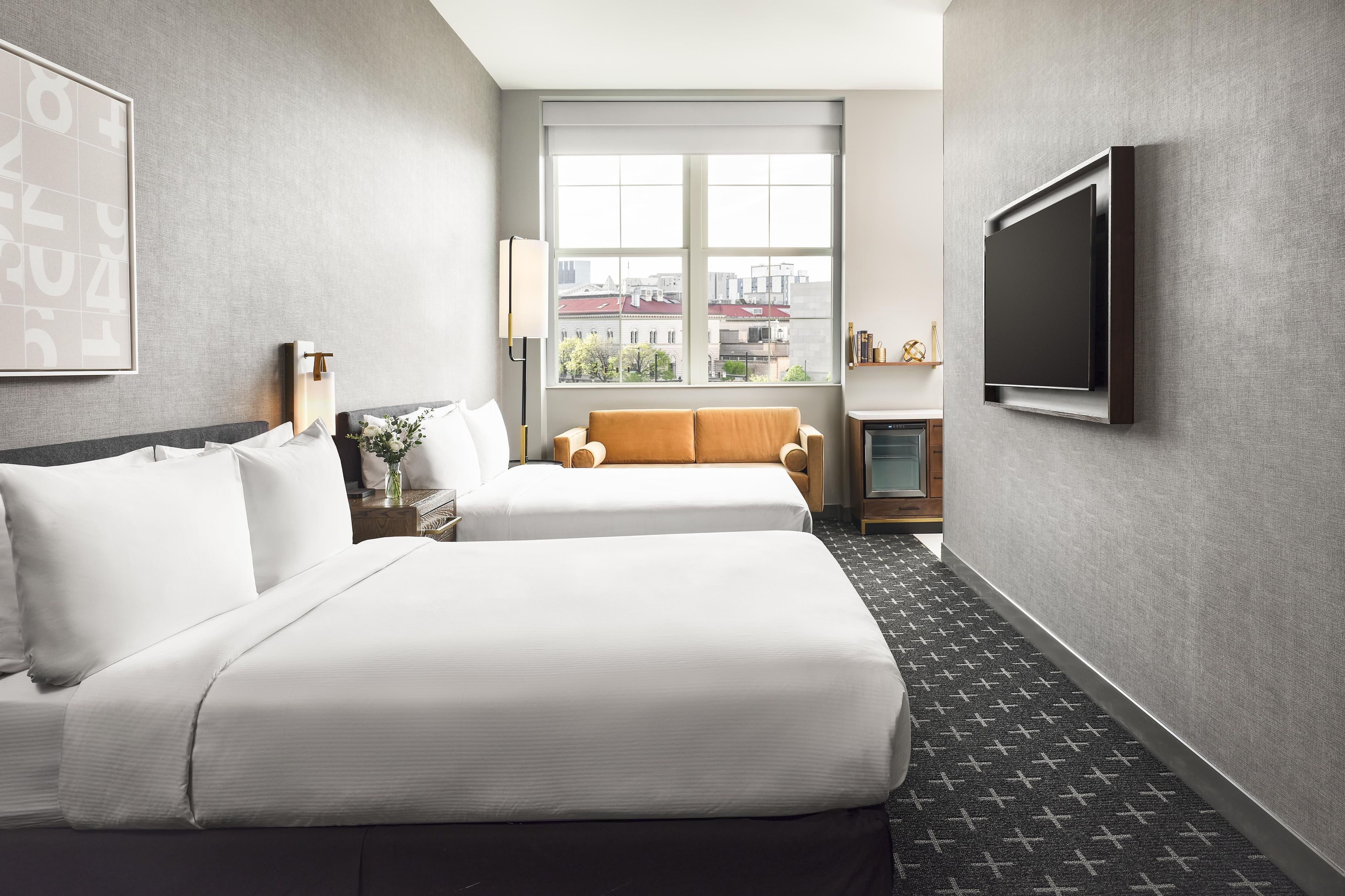 A hotel room with two beds, a large window with a city view, a flat-screen TV, and a small table with a lamp.