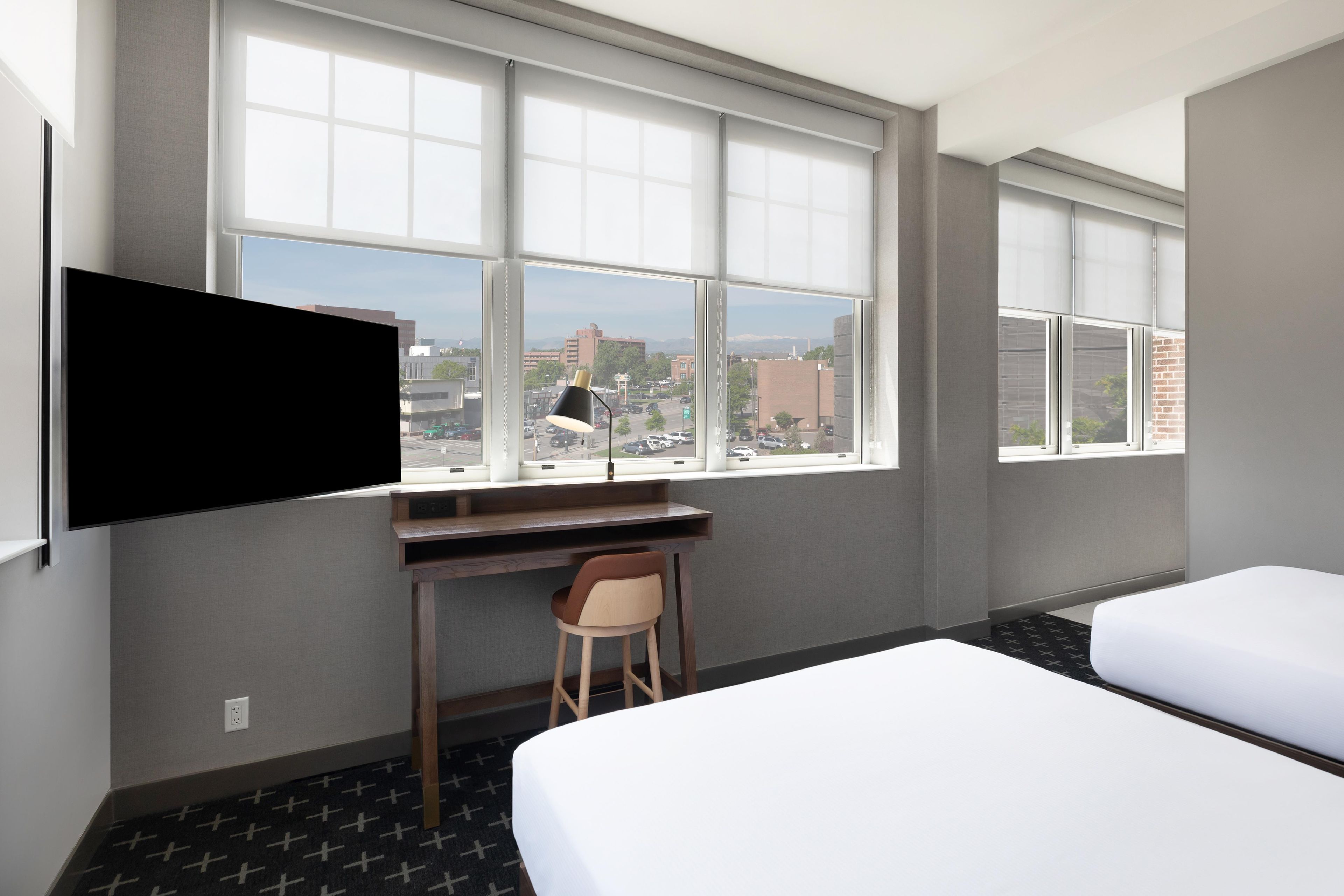 A hotel room with a bed, a desk, a chair, a TV, and large windows with a city view.