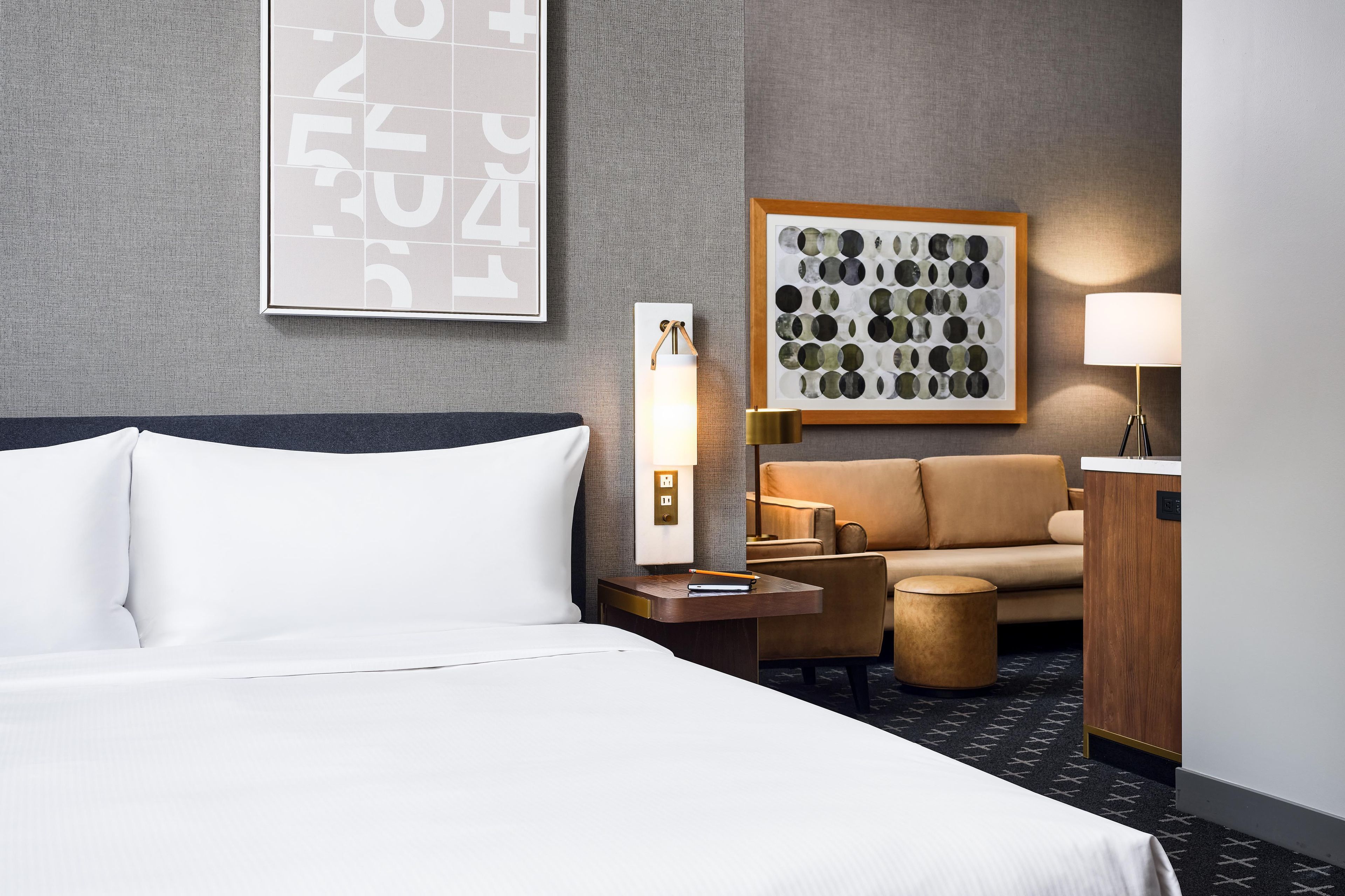 A hotel room with a bed, a nightstand, a lamp, a sofa, and a framed artwork on the wall.