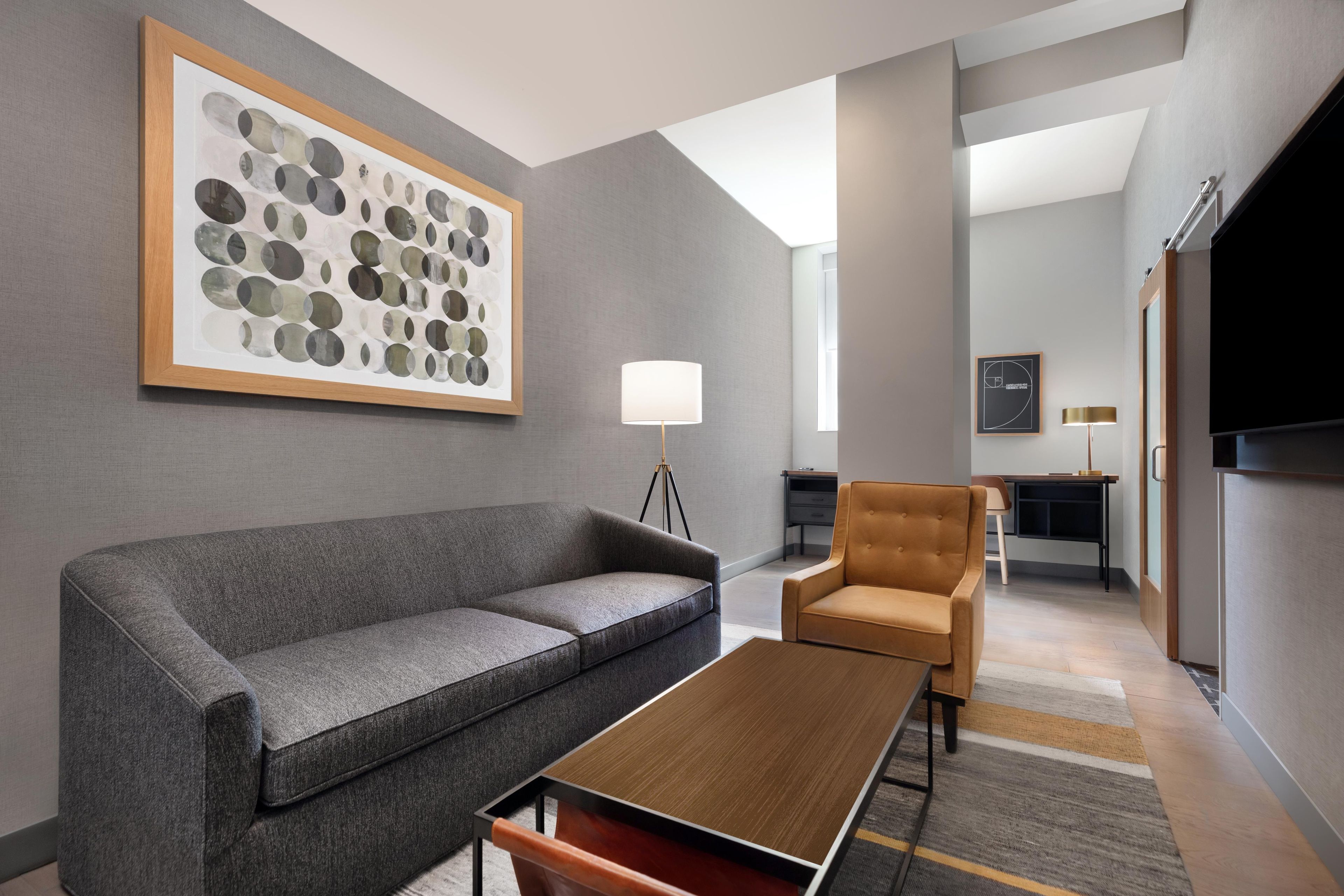A modern hotel room with a grey sofa, a wooden coffee table, a brown armchair, and a large abstract painting on the wall.