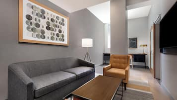 A modern hotel room with a grey sofa, a wooden coffee table, a brown armchair, and a large abstract painting on the wall.