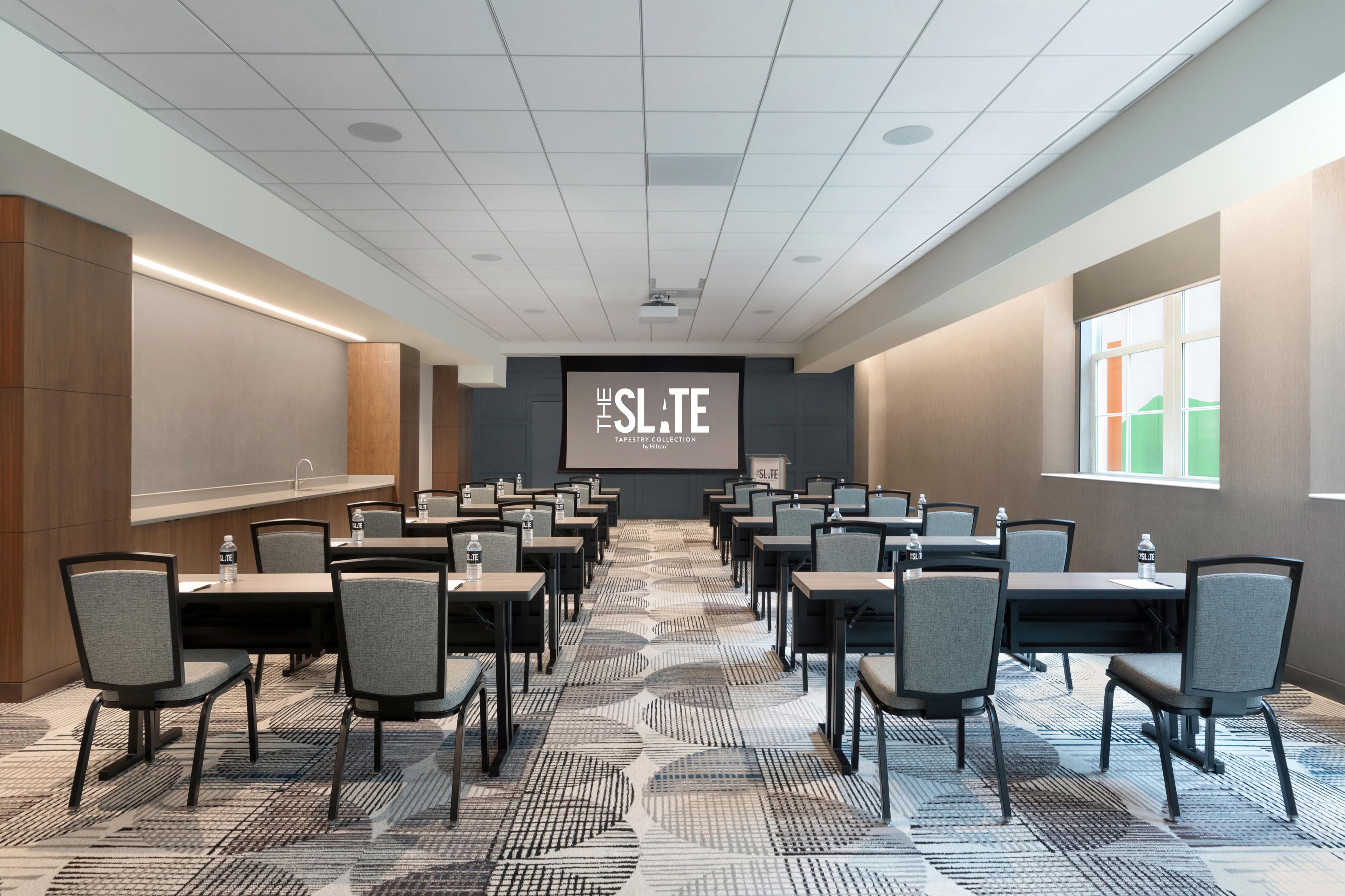 Un salón de conferencias con una pantalla para presentaciones, varias mesas y sillas dispuestas para reuniones.