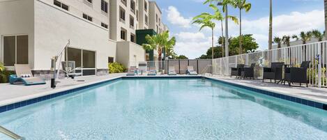A swimming pool with lounge chairs and palm trees, surrounded by a building with multiple windows and balconies.