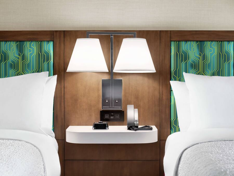 A hotel room with two beds, a bedside table with a lamp, a clock, and a phone.