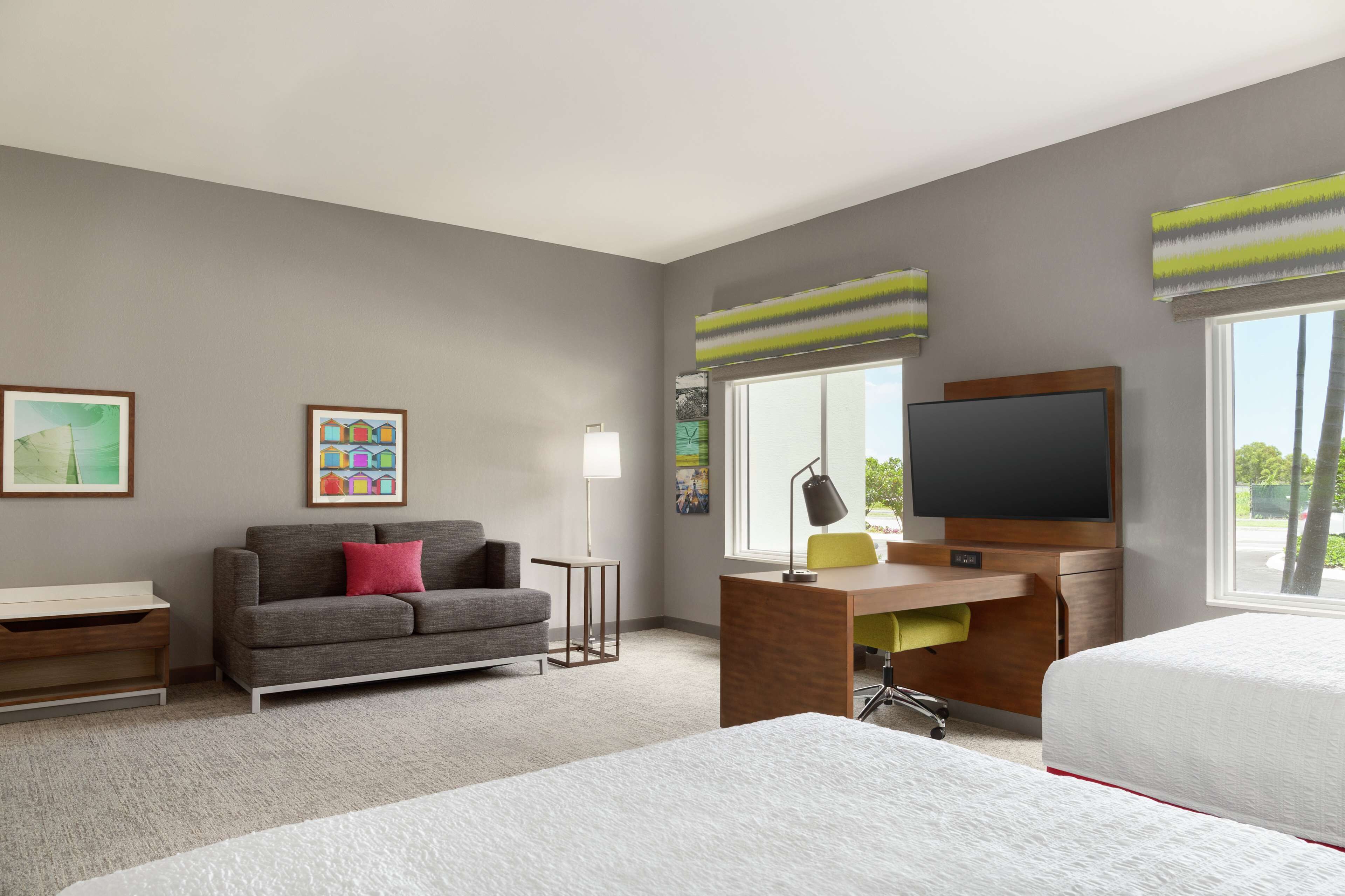 A hotel room with a grey sofa, a desk with a computer, a TV, and two beds.