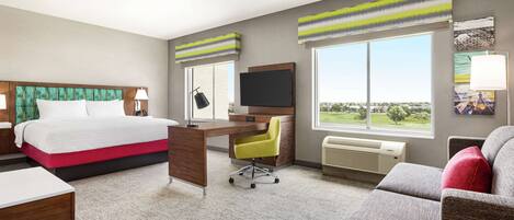 Habitación de hotel con cama, escritorio, silla, sofá y ventanas amplias.