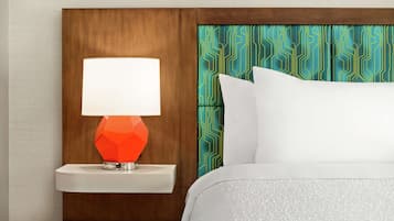 Quarto de hotel com cabeceira de madeira, mesa de cabeceira branca e luminária laranja geométrica, além de cama com roupa de cama branca.