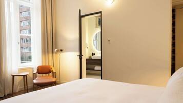 Une chambre d’hôtel avec un lit, une chaise, un bureau, un miroir et une fenêtre avec des rideaux.