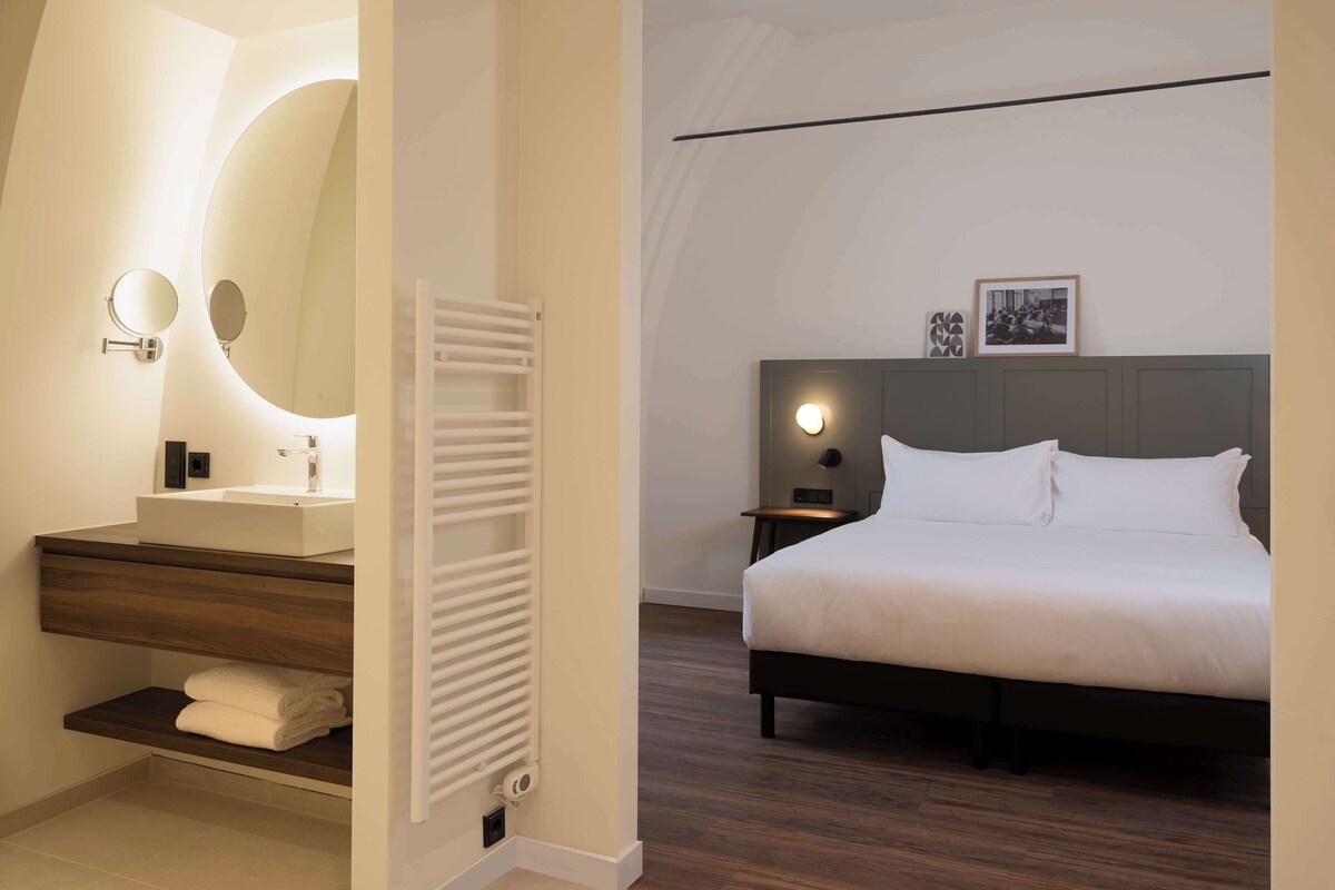 Habitación de hotel moderna con una cama grande, una mesita de noche con lámpara, un espejo redondo, un estante empotrado con toallas y un radiador blanco.