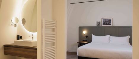 A modern hotel room with a large bed, a bedside table with a lamp, a round mirror, a built-in shelf with towels, and a white radiator.